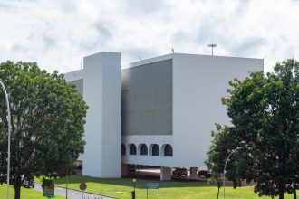 Edifício público branco de arquitetura moderna cercado por vegetação sob céu azul.
