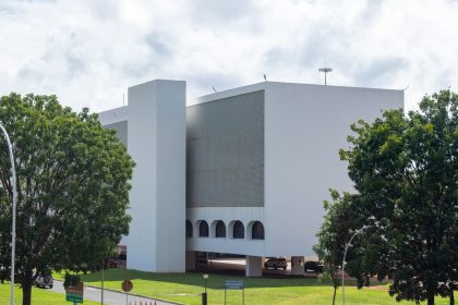 Edifício público branco de arquitetura moderna cercado por vegetação sob céu azul.