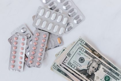 Blisters de medicamentos e comprimidos dispostos sobre uma pilha de cédulas, vistos de cima.