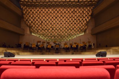 Interior de uma sala de concertos com palco iluminado e fileiras de assentos vazios, preparado para uma apresentação.