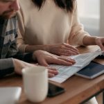 Homem e mulher analisam documentos financeiros à mesa, em casa, ao lado de um teclado e uma xícara de café.