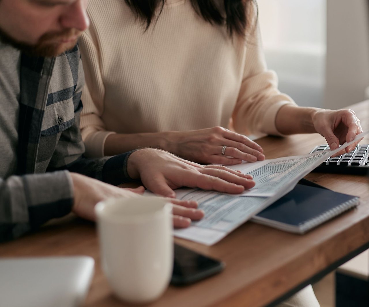 Homem e mulher analisam documentos financeiros à mesa, em casa, ao lado de um teclado e uma xícara de café.