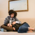 Mulher consola uma amiga que chora deitada em um sofá, em ambiente doméstico.