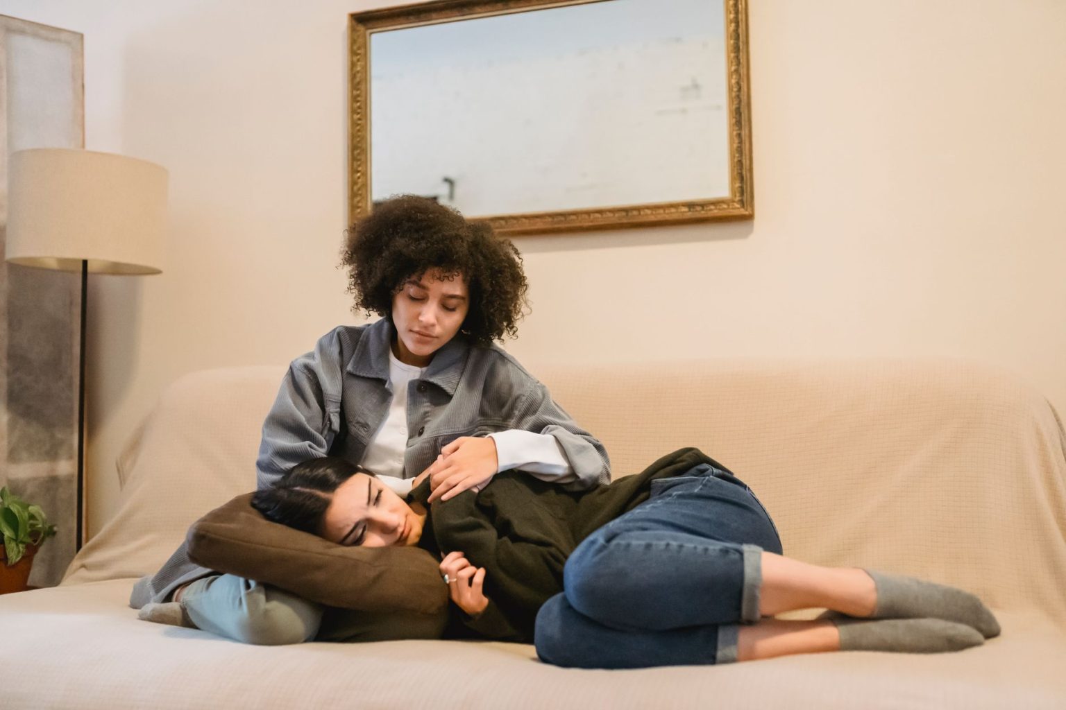 Mulher consola uma amiga que chora deitada em um sofá, em ambiente doméstico.