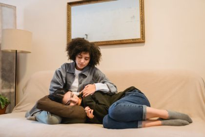 Mulher consola uma amiga que chora deitada em um sofá, em ambiente doméstico.