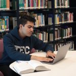 Estudante usa laptop e livros para estudar e realizar pesquisas em uma biblioteca universitária.
