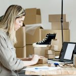 Empreendedora embala caixas para sua loja on-line, cercada por materiais de embalagem e um laptop.