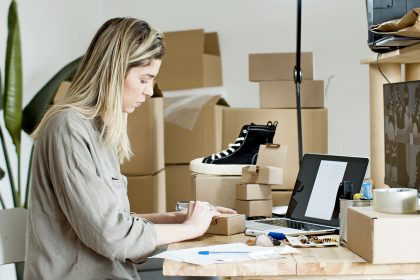 Empreendedora embala caixas para sua loja on-line, cercada por materiais de embalagem e um laptop.